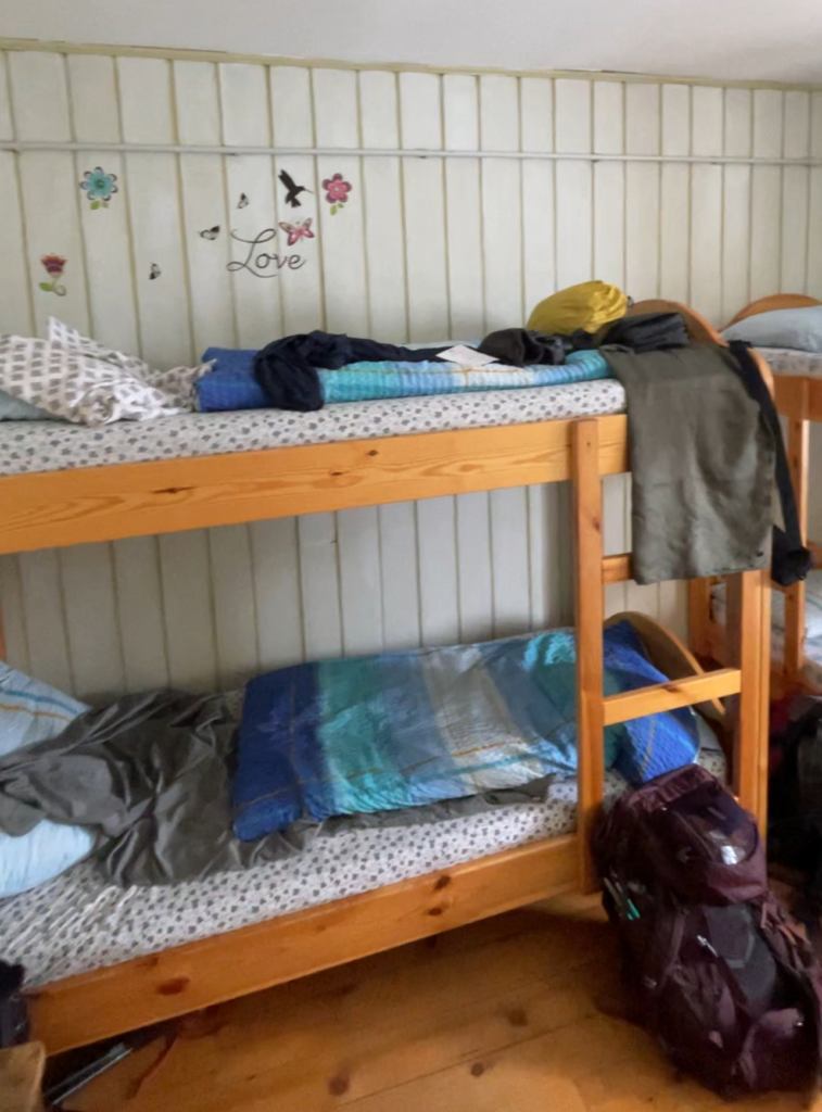 Wooden bunk bed setup in Rifugio Rosetta, with simple patterned bedding and hikers’ gear placed nearby. The white-paneled wall is decorated with flower and butterfly stickers and the word "Love," adding a cozy, personal touch to the shared dorm room.