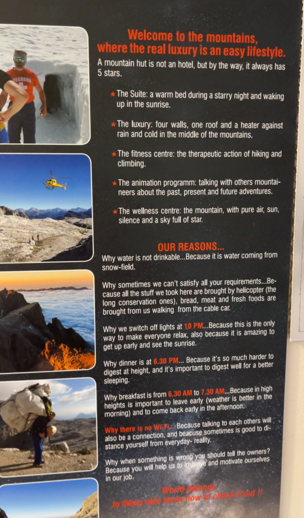 Informational sign at a Dolomites mountain hut with the title "Welcome to the mountains, where the real luxury is an easy lifestyle," humorously outlining the unique “luxuries” of hut life, such as a warm bed, shelter from rain, and hiking as a fitness center. It explains why there’s no Wi-Fi, why water isn’t drinkable, and details about dinner and breakfast times, quiet hours, and supply logistics, emphasizing simplicity, connection, and mountain culture.