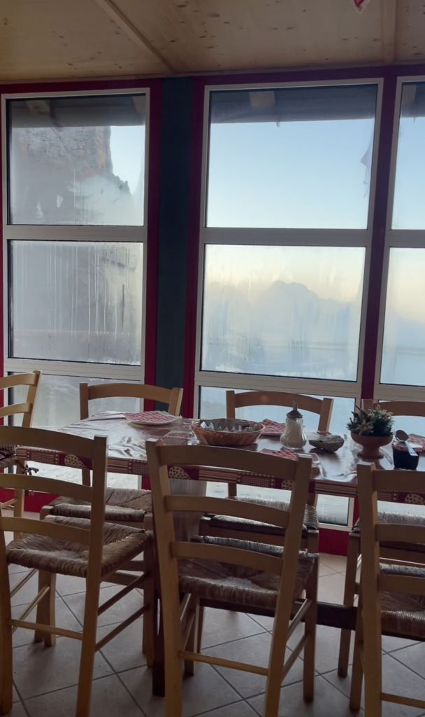 Cozy dining room at Rifugio Pradidali, featuring a wooden table set with breakfast items and surrounded by simple wooden chairs. Large fogged windows reveal a faint outline of Dolomite peaks in the early morning light.