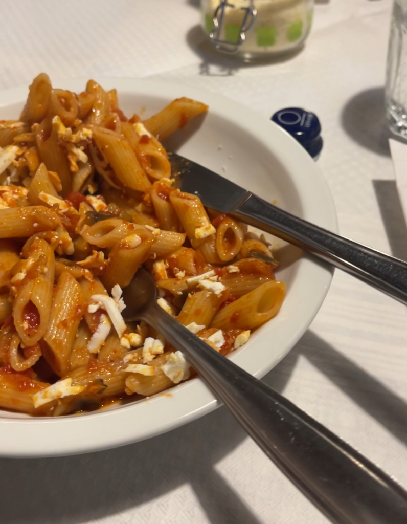 Bowl of penne pasta in tomato sauce served at a Rifugio, topped with crumbled cheese and visible bits of vegetables like mushrooms. A spoon and knife rest in the bowl on a white tablecloth, with a glass and grated cheese jar in the background.