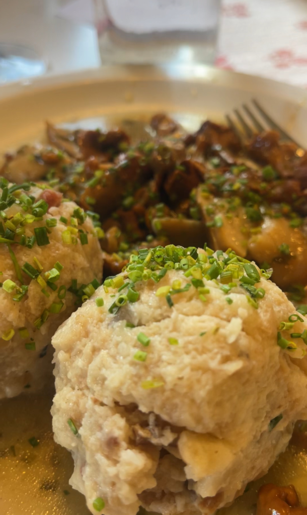 Traditional Tyrolean bread dumplings topped with fresh chives, served at Rifugio Genova/Schlüterhütte. The dumplings are paired with a savory mushroom sauce, offering a hearty alpine meal on a shallow plate.