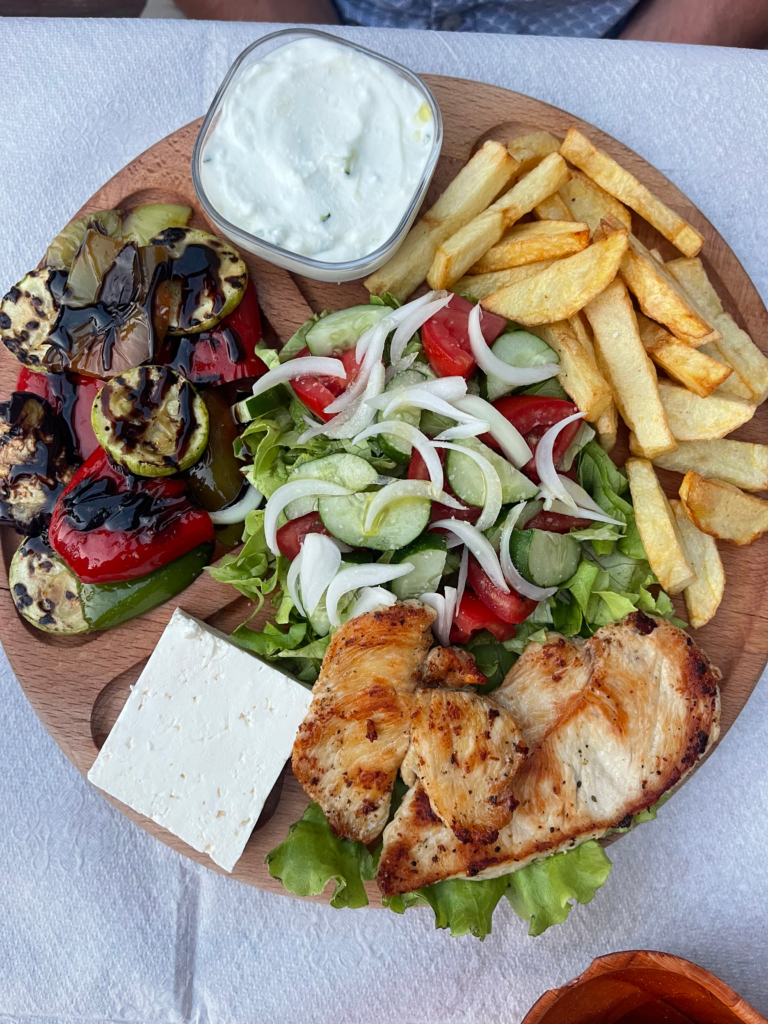 A display of food on a wooden cutting board with grilled vegetables, chicken, fries and a balkan salad at a guesthouse in Theth