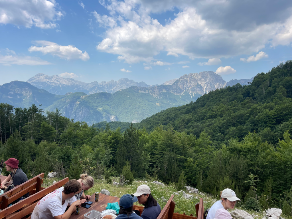 onlookers sitting at mountain hut and picnic tables with the view of the Albanian Alps