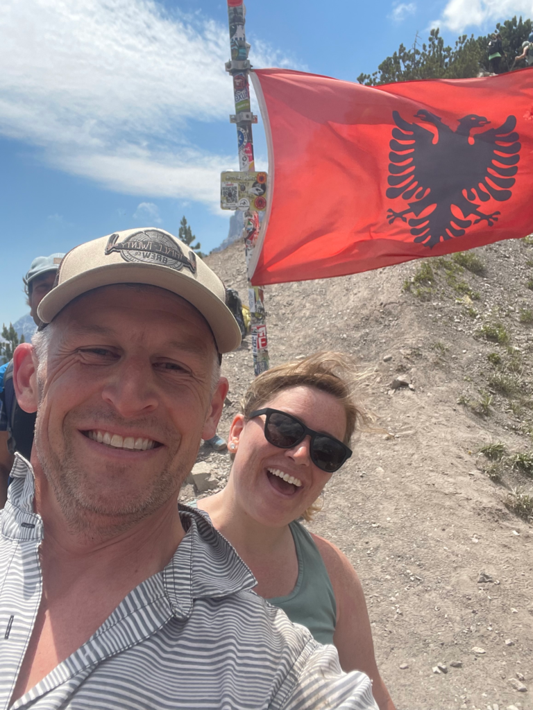 man and woman smiling at the camera as the the red and black albanian flag waves at the pass of valbone