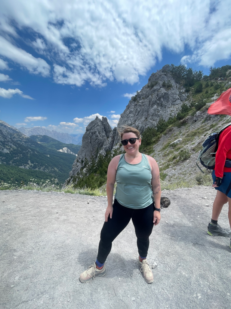 woman in a green tank top, black pants and sunglasses on standing at a mountain pass in the Albanian Alps on the route to Valbone Valley