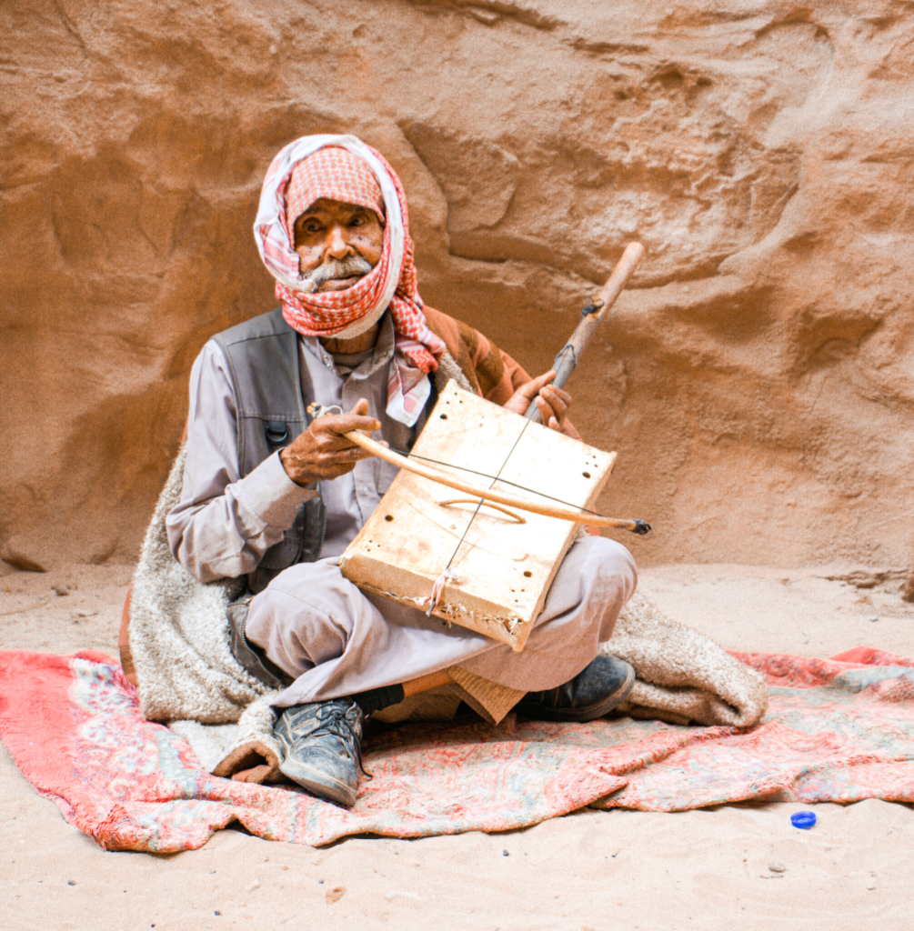 a bedouin man with his rababa sitting on the ground on a red blanket in Little Petra, Jordan