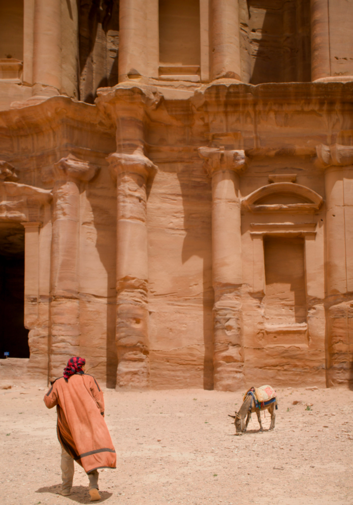 a man in desert clothing walks toward the monastery in Jordan with a donkey in the background.