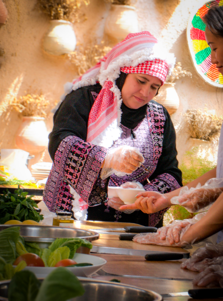 a woman in a red and white keffiyah helps a group prepare maqluba at beit khayrat souf