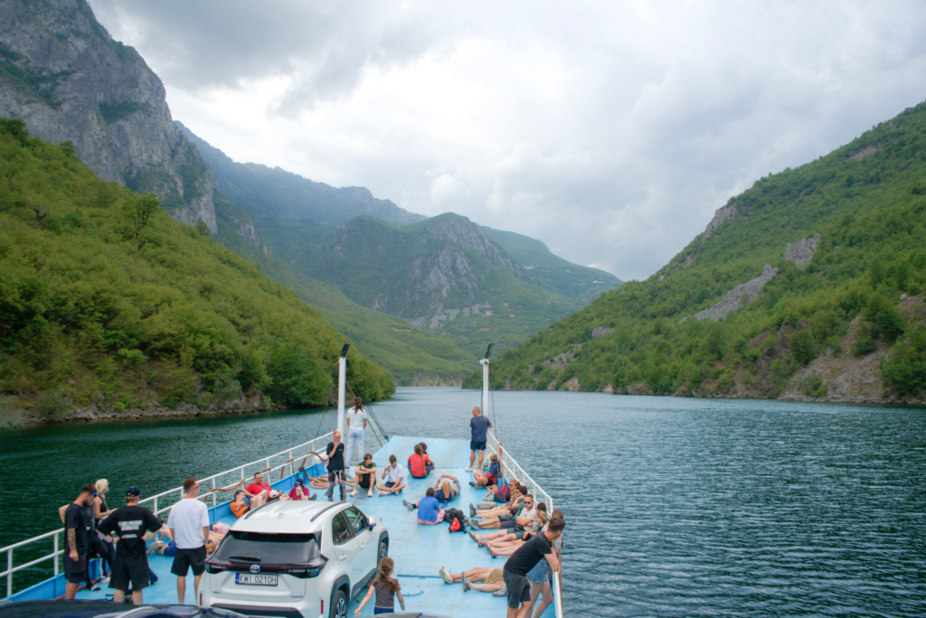 A view of the front side of the boat with people at the front and komani lake surrounding them with green albanian mountains to the side on a cloudy day