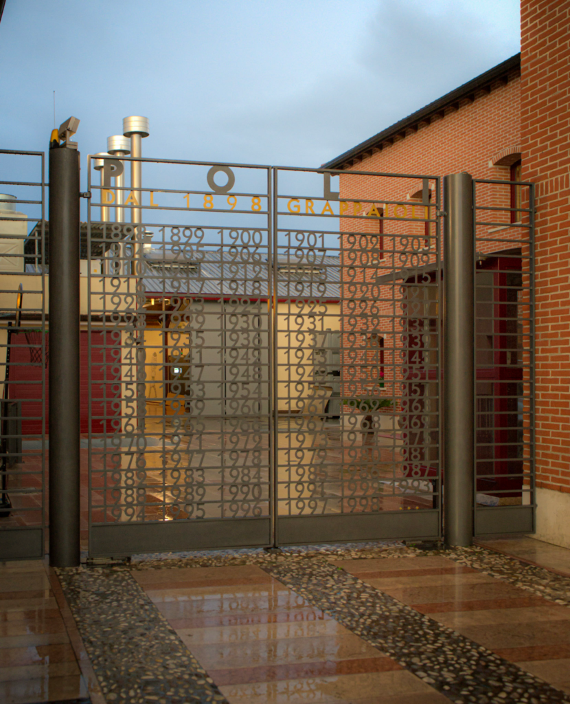 Gate of the Poli Grappa Distillery that is wrought iron with an orange and red brick building behind it