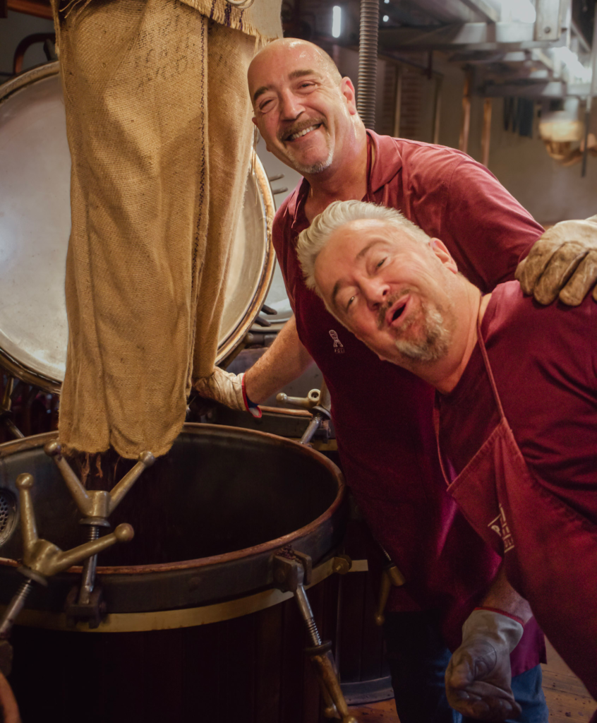 Two men smile at the camera next to a grape skin extraction barrel wearing red shirts that say Poli Grappa