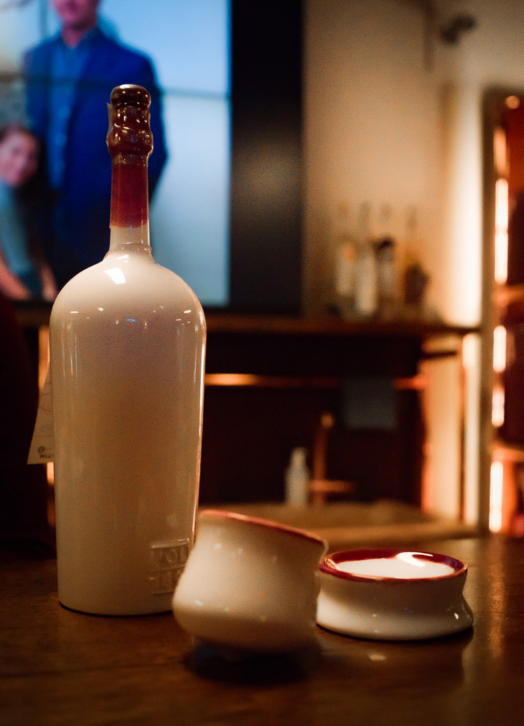 Traditional rasentin set at the Poli Grappa Museum and Shop in Schiavon Italy featuring a white ceramic bottle with a dark red glazed neck and matching small ceramic cups filled with clear grappa. The set is displayed on a wooden table in a warmly lit tasting room, highlighting Italian grappa culture and regional serving traditions.
