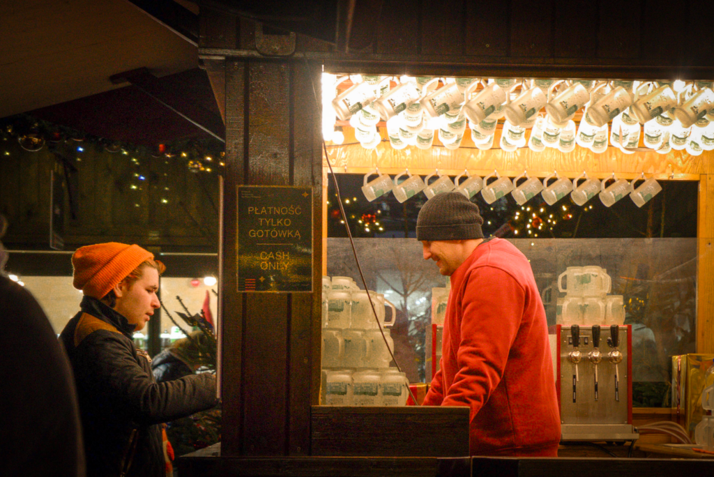 a man in a red sweater with a black beanie stands in a Christmas Market stall in Poznan talking to a customer with a cup in his hand