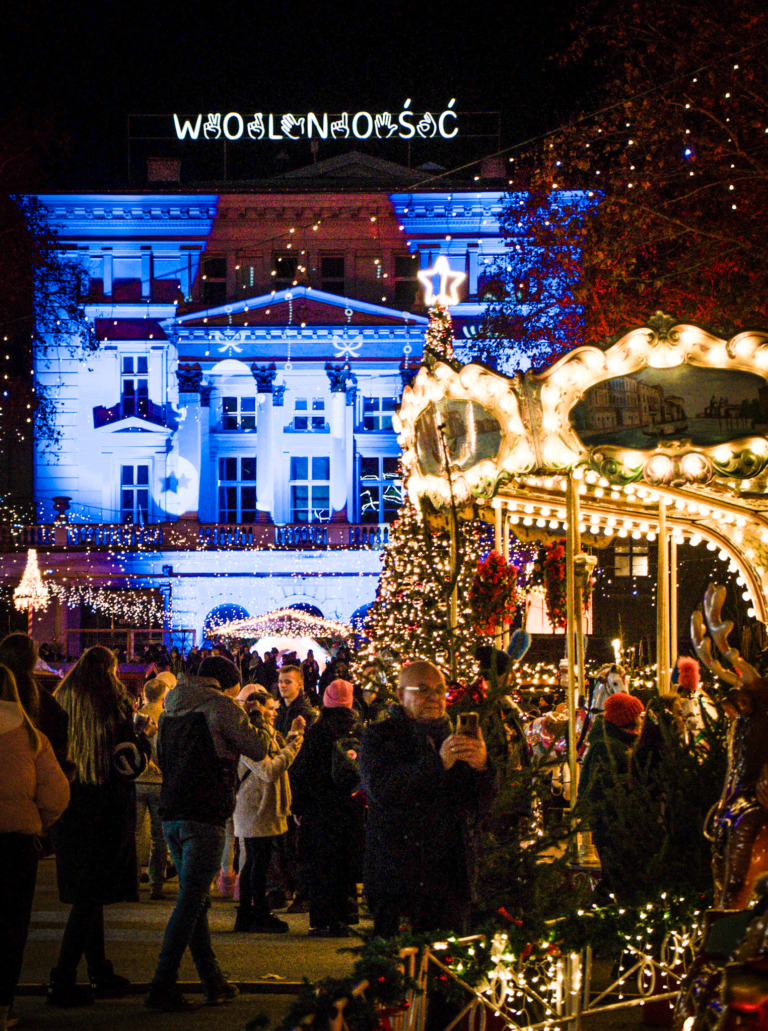 A neoclassical building illuminated with blue and red lights features the Polish word “Wolność” meaning “freedom” on its rooftop. In front of it, crowds gather among Christmas trees and festive lights at the Poznan Christmas Market.