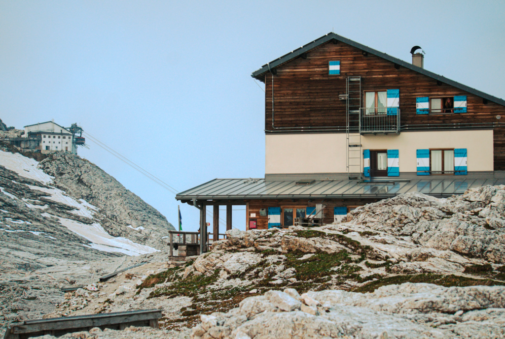 Mountain rifugio with blue shutters perched on rocky terrain in the Dolomites with patches of snow and a cable car station visible in the background. The alpine hut sits along a high elevation trail offering a scenic stop for hikers seeking one of the best rifugios in the Dolomites on an easy day hike.