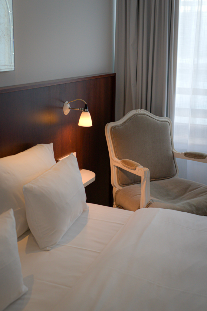 the interior of a hotel room with a white bed, dark brown wood headboard and grey clothed chair next to it at Ruby Coco Hotel