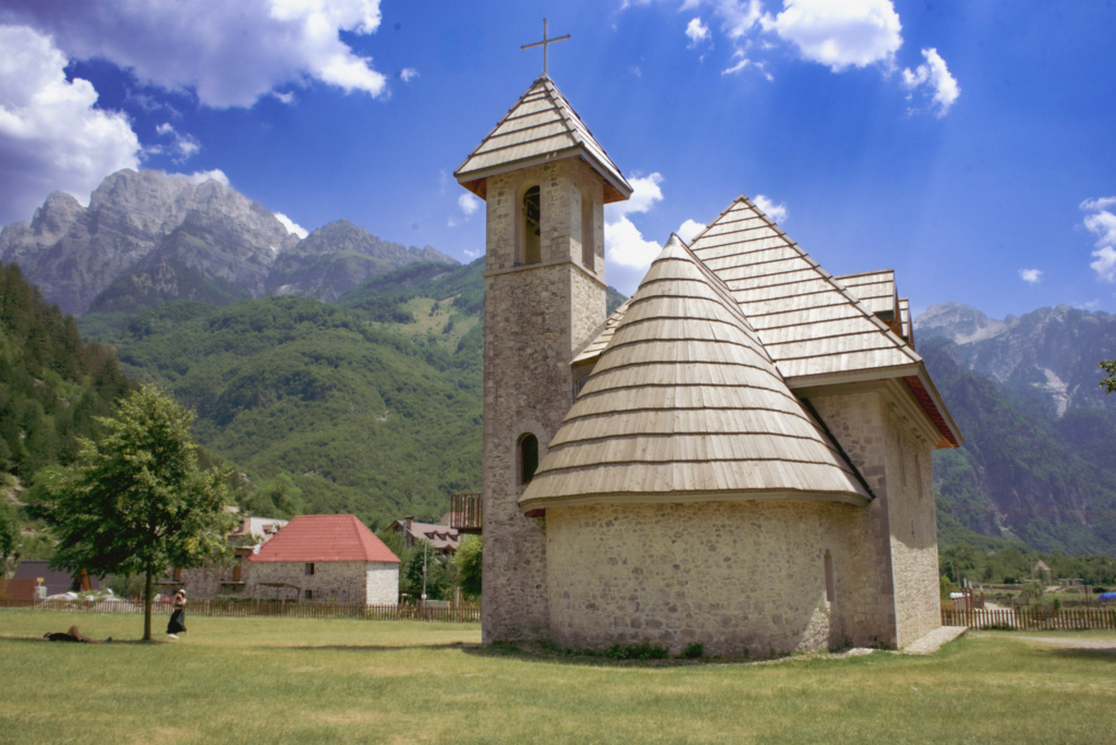 Stone church with a wooden roof and bell tower set in a lush green valley in Theth, Albania, with dramatic mountain peaks in the background under a blue sky. A hiker rests under a tree, capturing the serene atmosphere of the Albanian Alps.