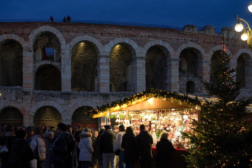 a lit up christmas market stall with passerbys and a christmas tree while the verona arena is in the background