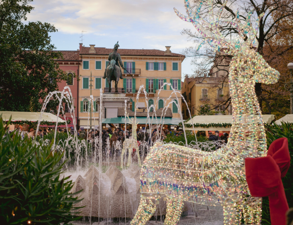 A Colorful reigndeer with a red bow and a statute in the back while a Fontana Bra goes off in Piazza Bra