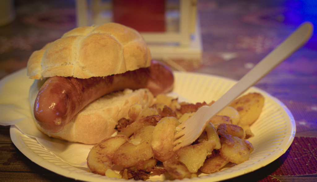 wurstel on a white bread bun with fried potatoes and a fork stuck in them at the Verona Christmas Market