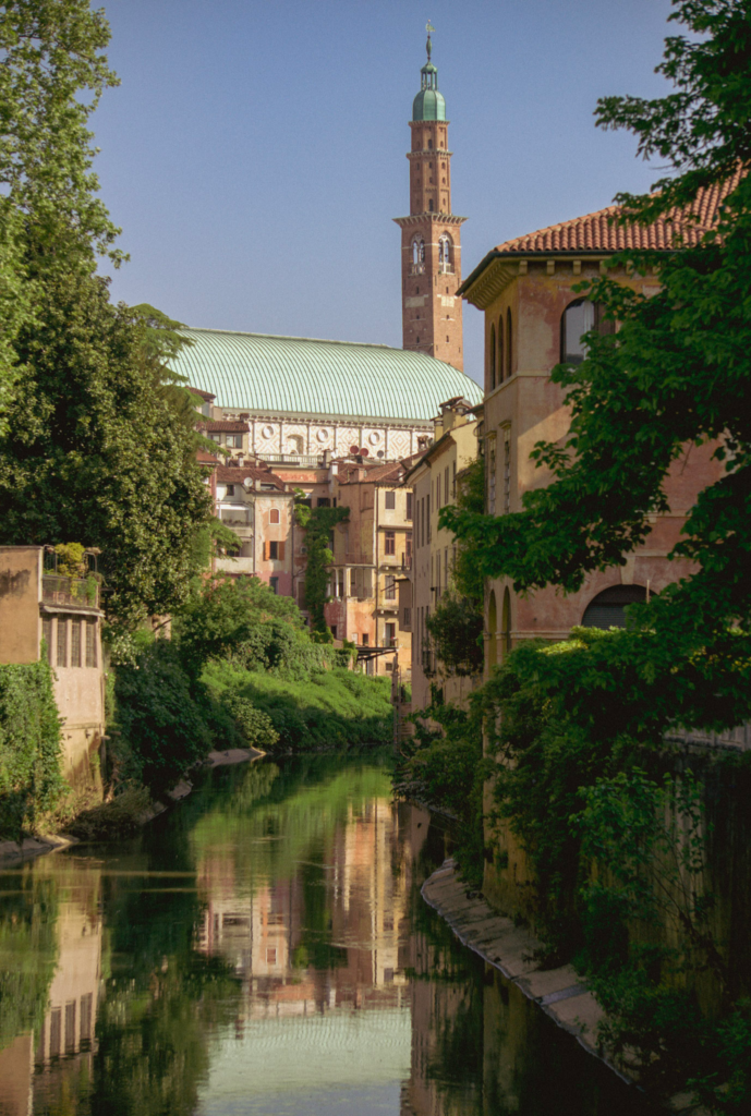 a view of the Fiume Retrone in Vicenza, Italy with lush greenery on the riverside and the Basilica peeking out beyond the photo