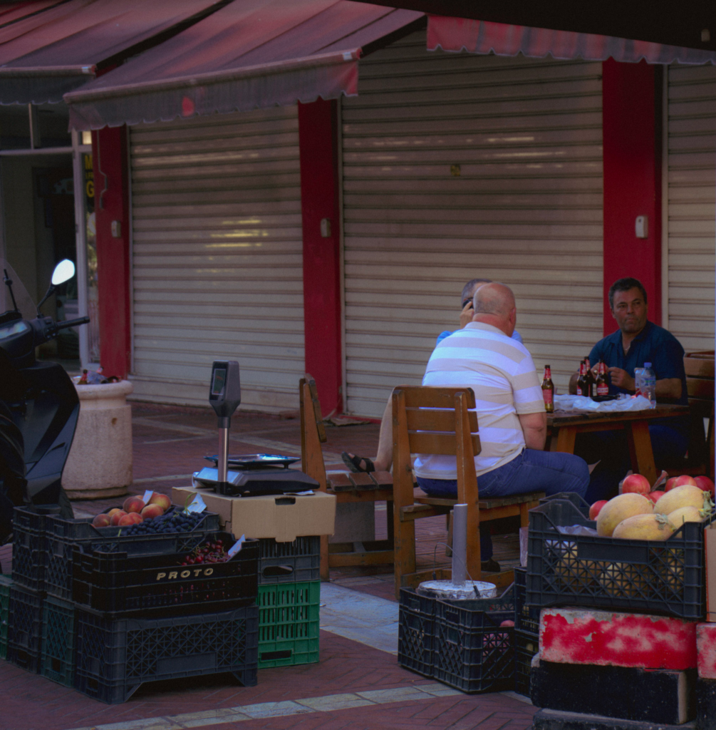 2 men sit at a table near a shop in Tiranas Piazza i Ri with boxes sitting around them