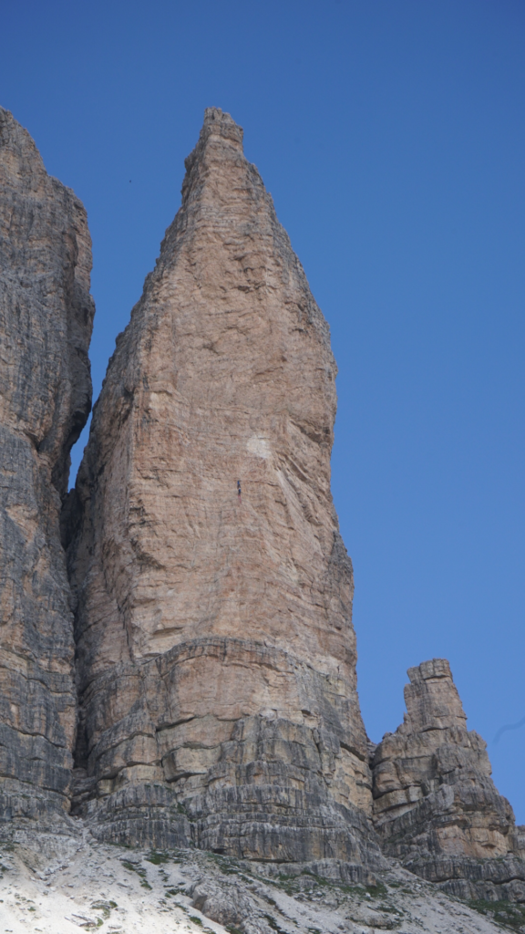 A towering pointed rock spire with a tiny climber halfway up its vertical face, captured against a clear blue sky during the Tre Cime di Lavaredo hike.