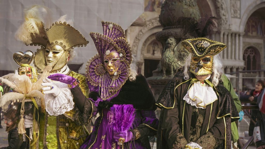Group of three people dressed in ornate 18th-century inspired costumes with tricorn hats and decorated masks, standing in front of the iconic domes and arches of Venice. The clothing features lace, brocade, and feathers, reflecting the historical elegance of the Carnevale.