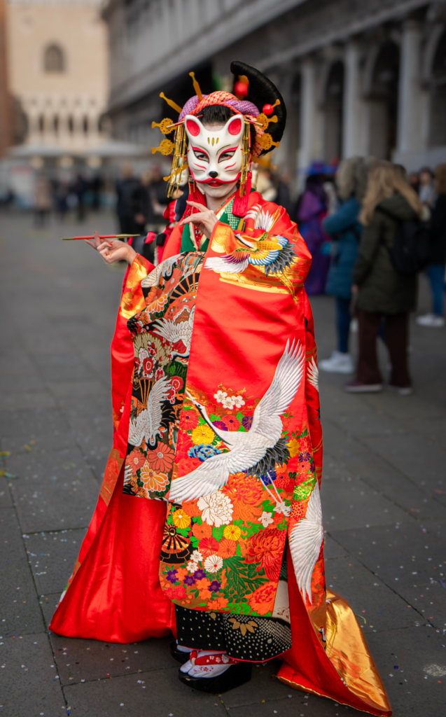 Individual in a striking red and gold kimono with floral and crane motifs, wearing a white fox mask and traditional-style hair ornaments, standing in a public square during Carnevale. The outfit blends Japanese cultural elements with the theatricality of the festival.