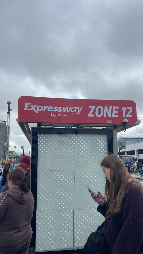 a woman standing next to a sign that is red and white that says Expressway Eirann Zone 12