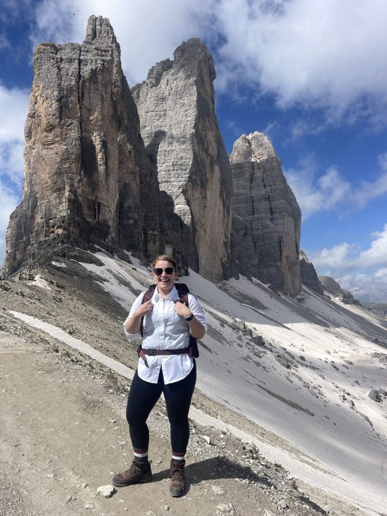 A smiling solo hiker stands on a rocky trail with trekking boots and a backpack, posing in front of the towering Tre Cime di Lavaredo peaks. The dramatic cliffs and snowy slopes highlight the epic scenery along the Tre Cime di Lavaredo hike.