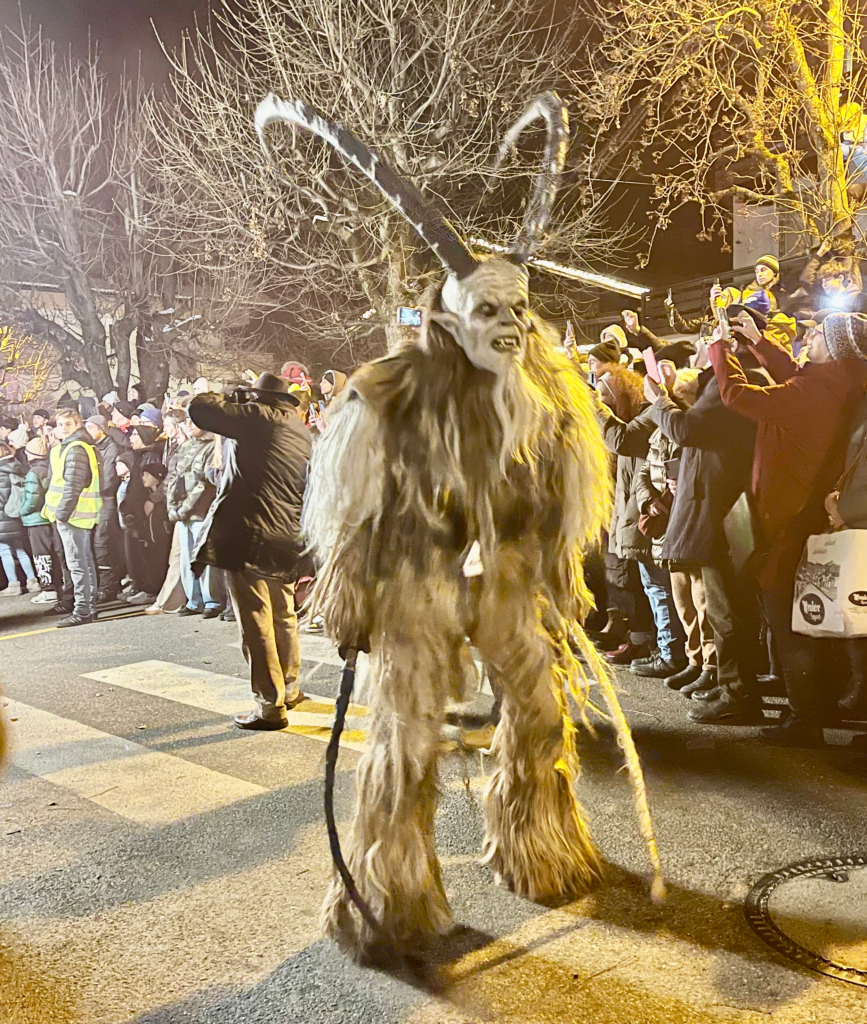 A Krampus character in a ghostly white and gray costume with long fur, oversized horns, and a snarling face approaches the camera, lit by yellow streetlamps. Crowds line the sidewalk under bare winter trees.