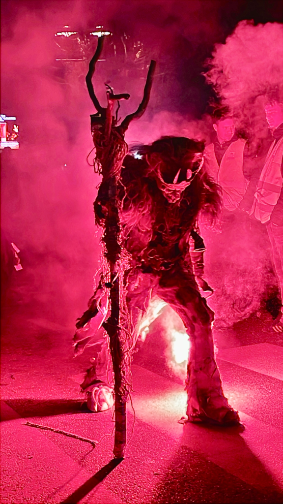 Two Krampus performers, barely visible in the heavy red smoke, walk side by side with lit torches and curved horns silhouetted against the haze.
