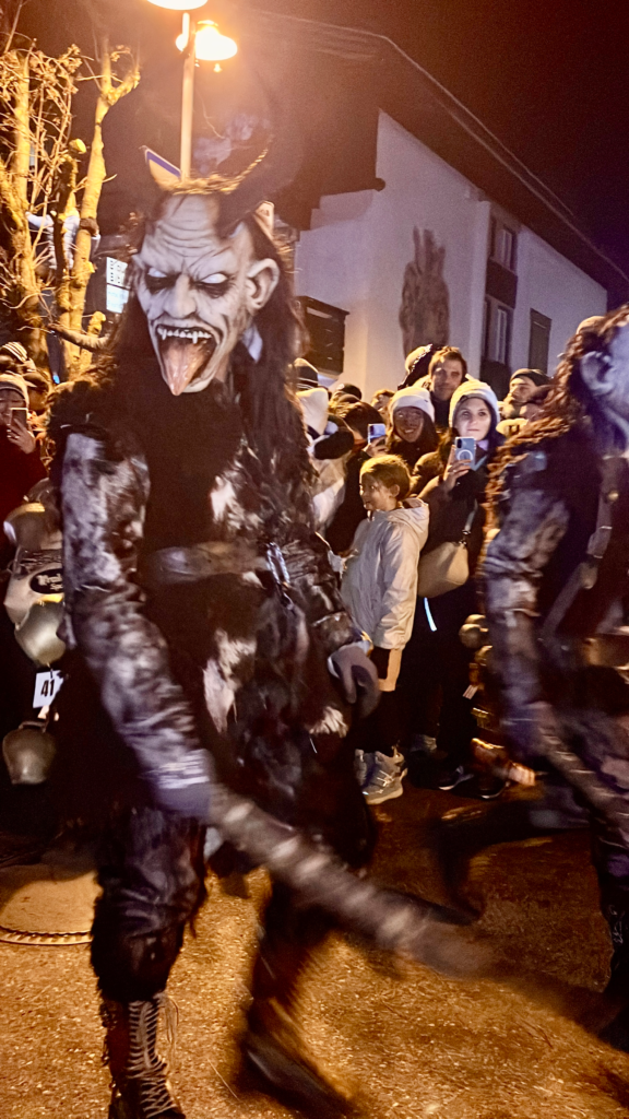 A fearsome Krampus with exaggerated facial features, a long tongue, and curled horns glares toward the camera. Crowds behind him appear captivated, some smiling and filming the moment.