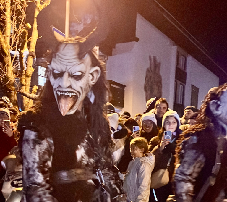 A fearsome Krampus with exaggerated facial features, a long tongue, and curled horns glares toward the camera. Crowds behind him appear captivated, some smiling and filming the moment.