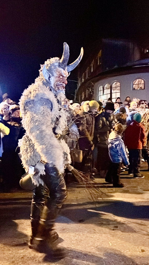 A white-furred Krampus character with menacing red eyes and twisted horns carries a bundle of sticks, walking past a winter crowd of adults and children at the Krampus parade.