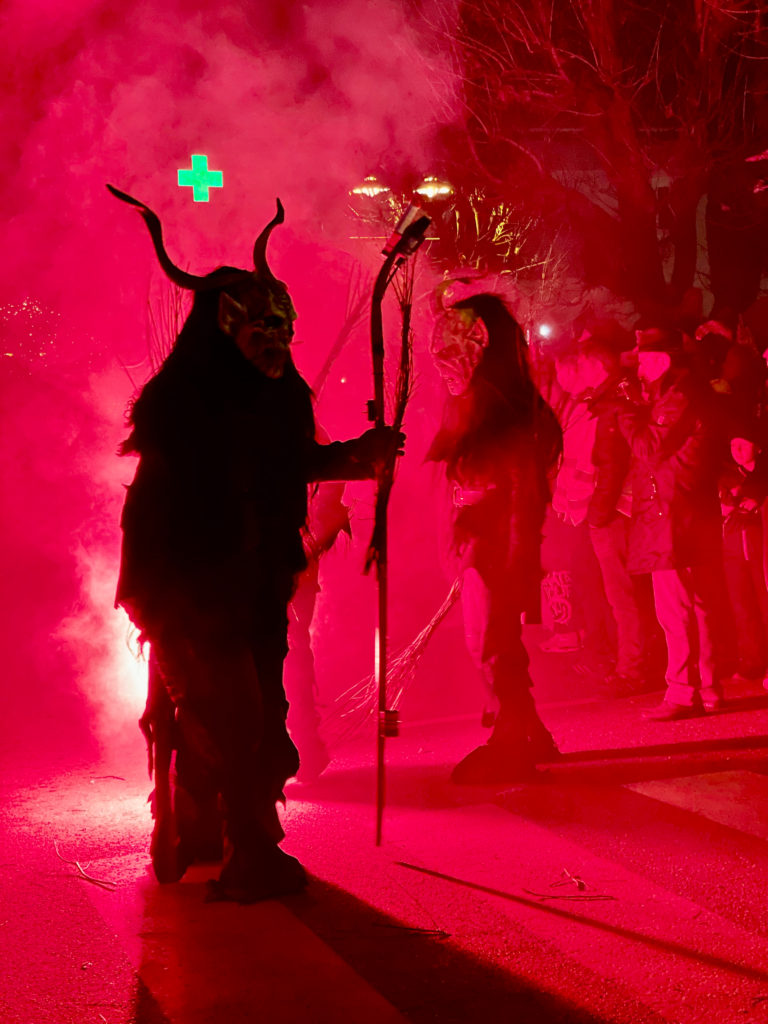 Silhouetted Krampus figures holding staffs or torches walk through thick red smoke during the nighttime parade. A glowing green pharmacy cross and crowds are faintly visible behind them.
