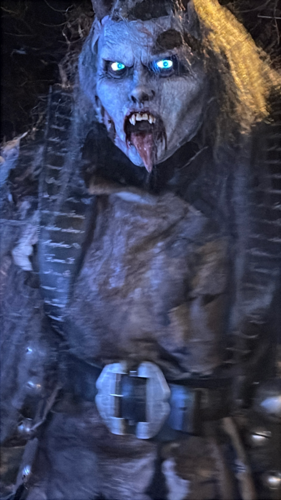 Close-up of a terrifying Krampus mask with glowing blue eyes, a bloodied mouth, and sharp fangs. The creature wears a dark fur costume with armor-like straps and a large metal belt buckle.