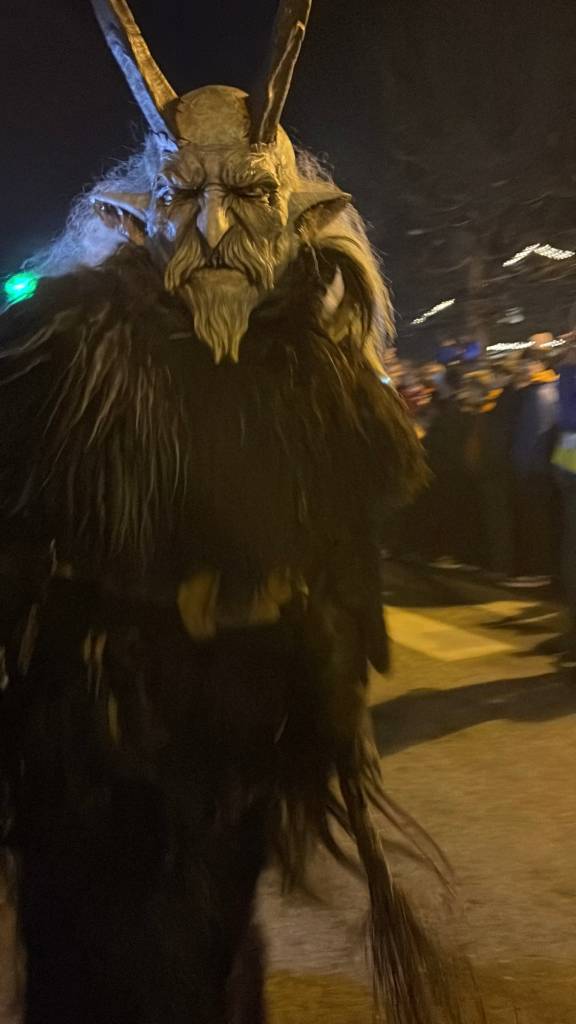 A Krampus with gnarled horns and a long white beard looms in the street during the Toblach Krampuslauf. Onlookers and security staff watch from the sides.