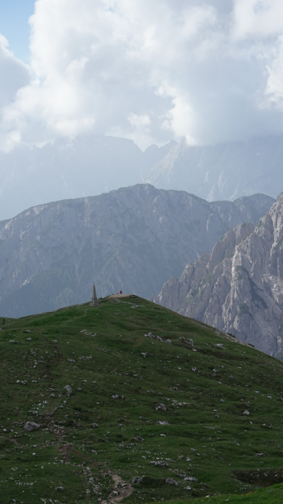 A steep grassy slope with a narrow trail leading to a stone obelisk monument on a ridge, with hazy gray cliffs and clouds in the background on the Tre Cime di Lavaredo circuit.