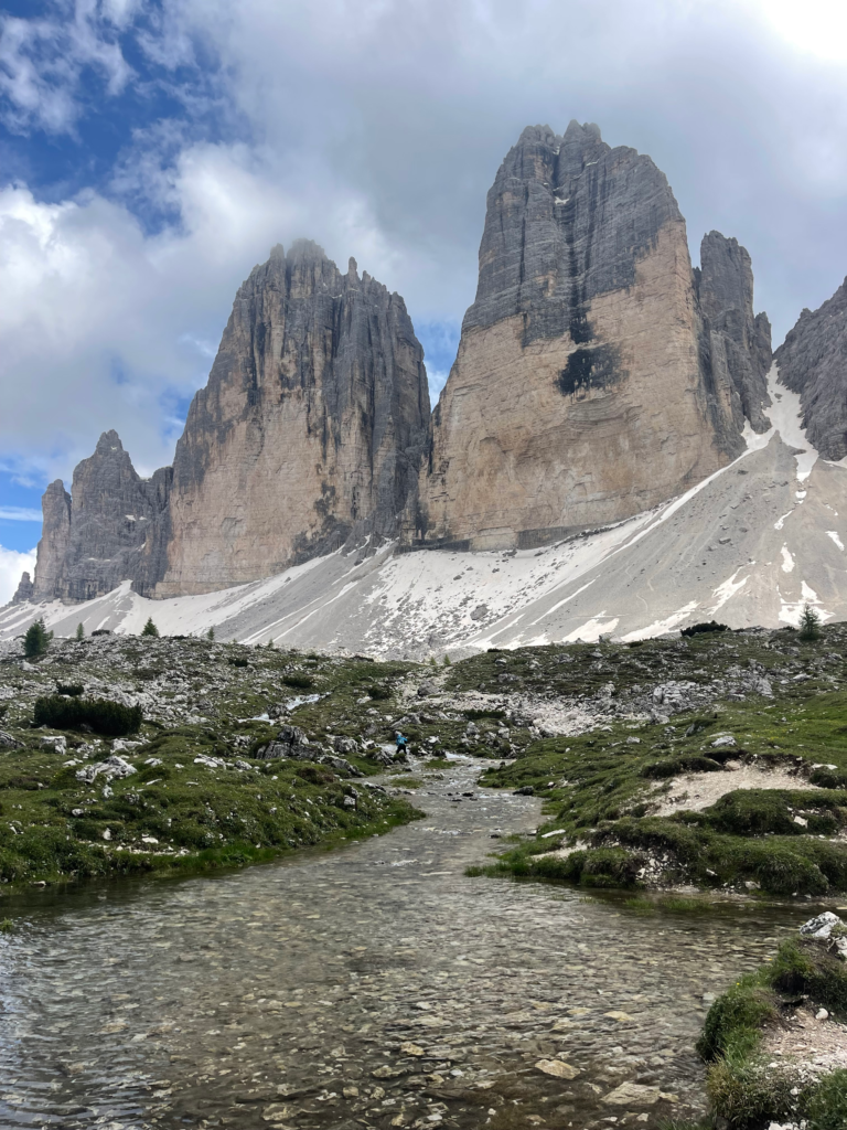 Two hikers walk along a grassy alpine trail beside a clear stream, approaching towering, jagged peaks dusted with snow on the Tre Cime di Lavaredo hike. The dramatic cliffs and cloudy sky add to the rugged mountain atmosphere.