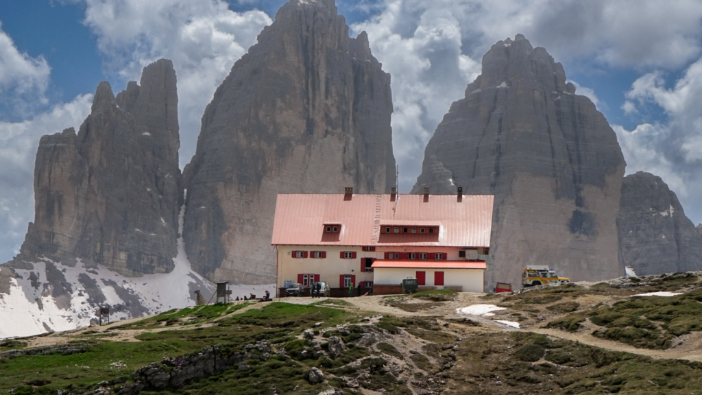 A mountain hut with a red roof and shuttered windows sits in the foreground of the towering Tre Cime di Lavaredo peaks. The dramatic rock formations and scattered snow patches highlight the rugged beauty of this iconic stop on the Tre Cime di Lavaredo hike.