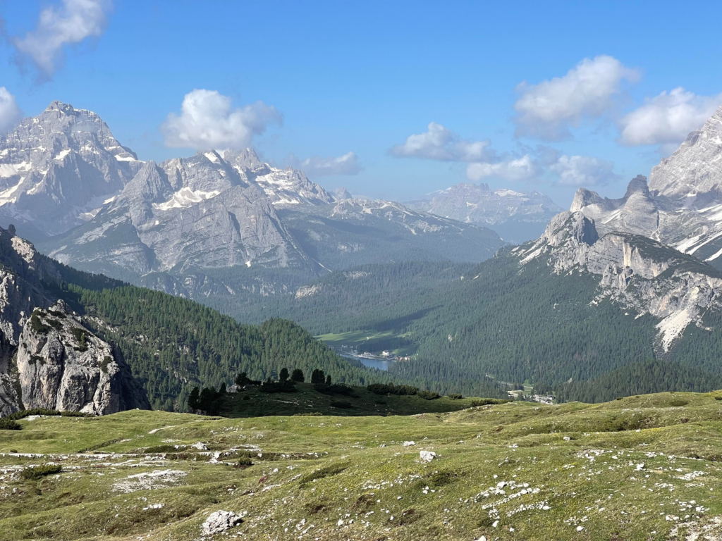 Wide-angle view of a lush alpine meadow with rocky trails leading toward the jagged peaks of the Dolomites under a vivid blue sky, seen during the Tre Cime di Lavaredo hike.