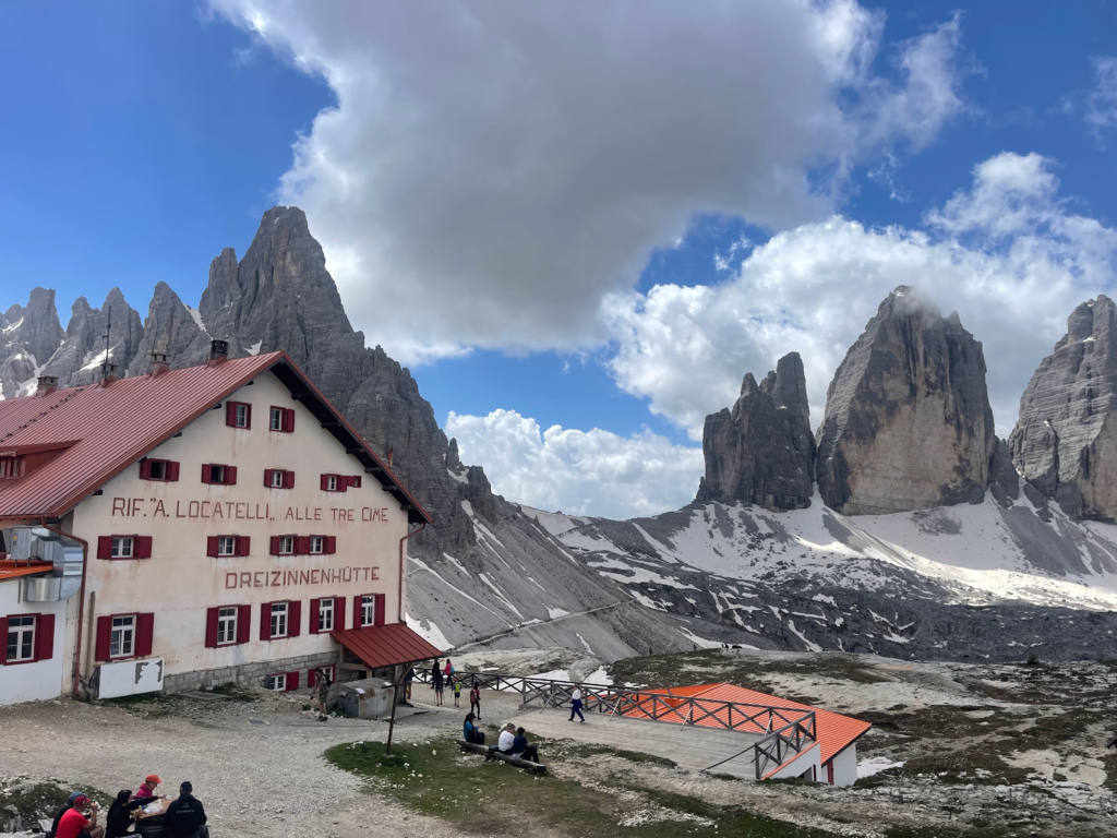 The Rifugio Locatelli mountain hut, with its red shutters and roof, stands at a scenic viewpoint facing the iconic peaks of Tre Cime di Lavaredo. Hikers rest nearby while dramatic limestone towers and snowy slopes stretch across the rugged landscape.