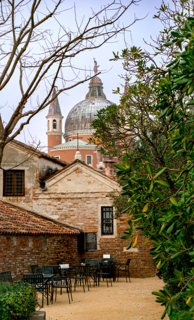 A peaceful courtyard with empty black metal tables and chairs set against rustic brick walls, framed by greenery and leafless tree branches, with the dome of the Church of the Most Holy Redeemer rising in the background. This serene corner captures the tranquil charm of the Convent Garden.