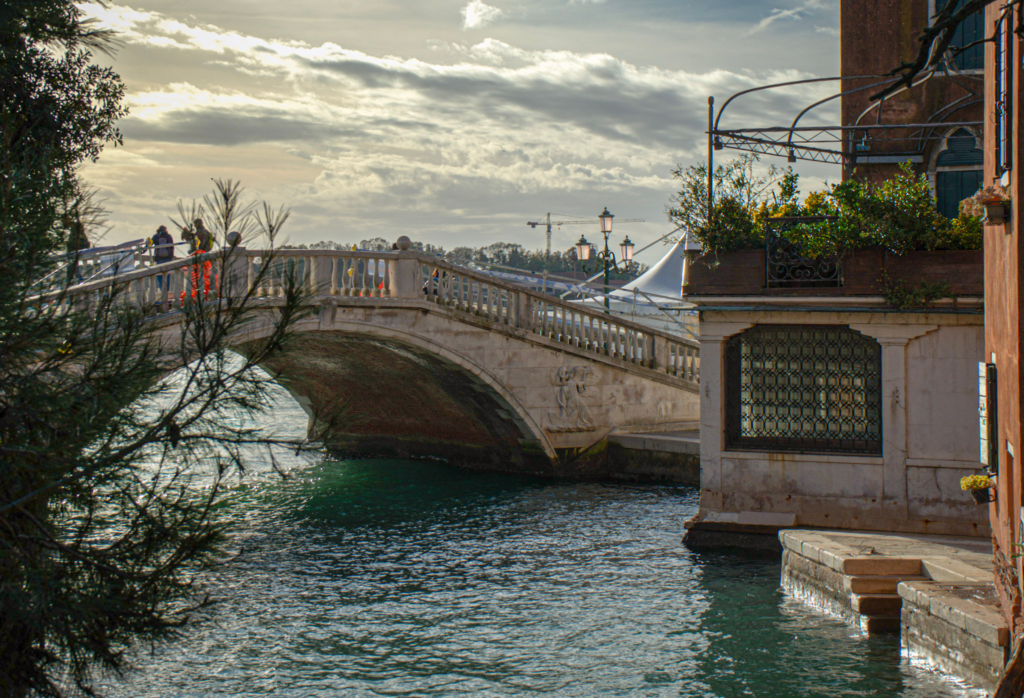 Stone bridge crossing a Venetian canal under a cloudy sky, with soft sunlight reflecting off the water and silhouetted pedestrians walking along the top. On the right, a historic building with wrought iron windows and a terrace garden overlooks the canal, capturing a quiet, scenic moment away from the crowds.