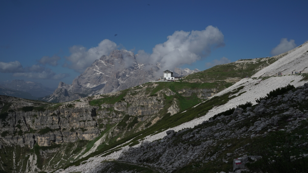 A distant view of a white mountain hut perched on a green ridge with towering rock faces behind it on the Tre Cime di Lavaredo hike. The dramatic Dolomites peaks rise into the clouds under a deep blue sky.