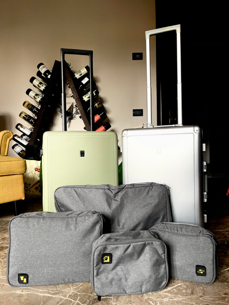 Five gray Level8 packing cubes in various sizes are arranged in front of two upright Level8 suitcases—one mint green and one silver. The cubes have icons indicating different packing uses, and the setup is indoors near a wine rack, illustrating travel organization options with Level8 luggage.