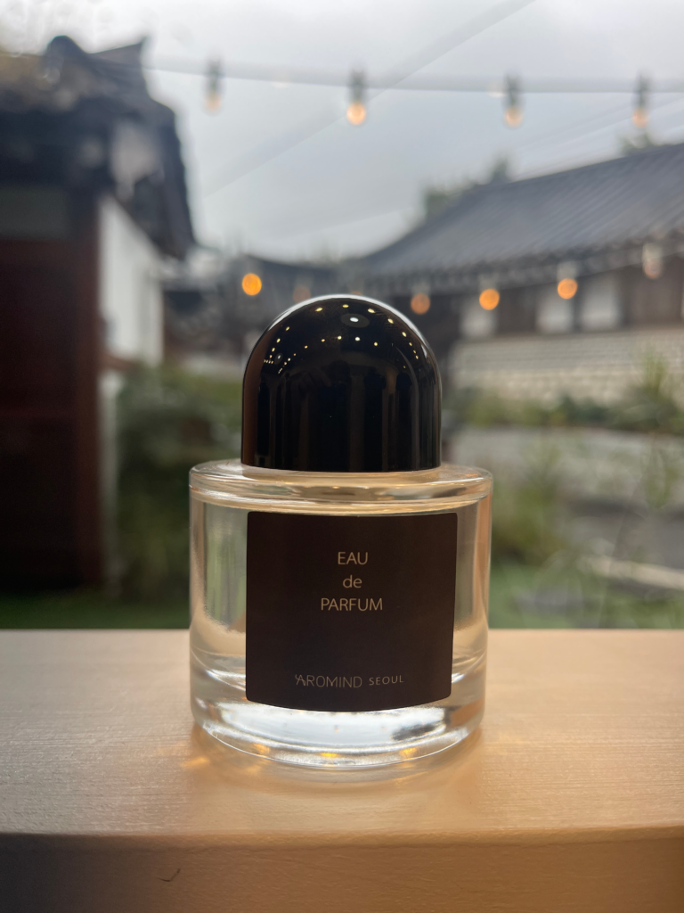 A bottle of Eau de Parfum from AROMIND Seoul sits on a windowsill with traditional Korean rooftops and string lights blurred in the background. Experiencing local fragrance design is an elegant thing to do in Seoul South Korea.