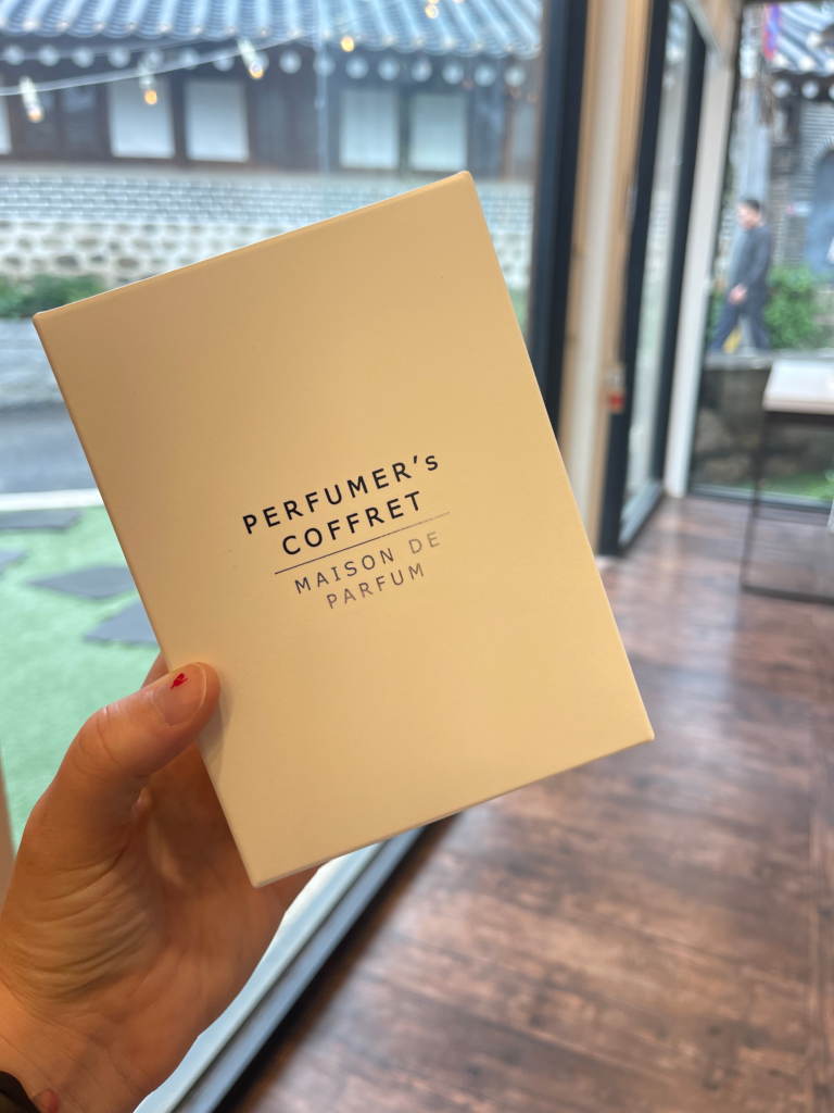 A hand holds a white box labeled “Perfumer’s Coffret Maison de Parfum” inside AROMIND, with a view of a hanok courtyard through the window. Participating in a custom perfume experience is a unique and sensory thing to do in Seoul South Korea. Let me know if you're also listing AROMIND specifically as a recommendation and want alt text adjusted to highlight that.